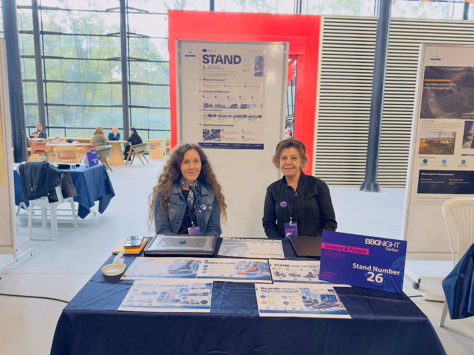 Two members of the ITU team seated at the STAND exhibition booth at BBGNight, with a STAND project poster on Smart Textile Diabetic Foot Care displayed behind them. The table in front holds printed information sheets in Turkish and a purple BBGNight "Science & Future Night" sign showing Stand Number 26.